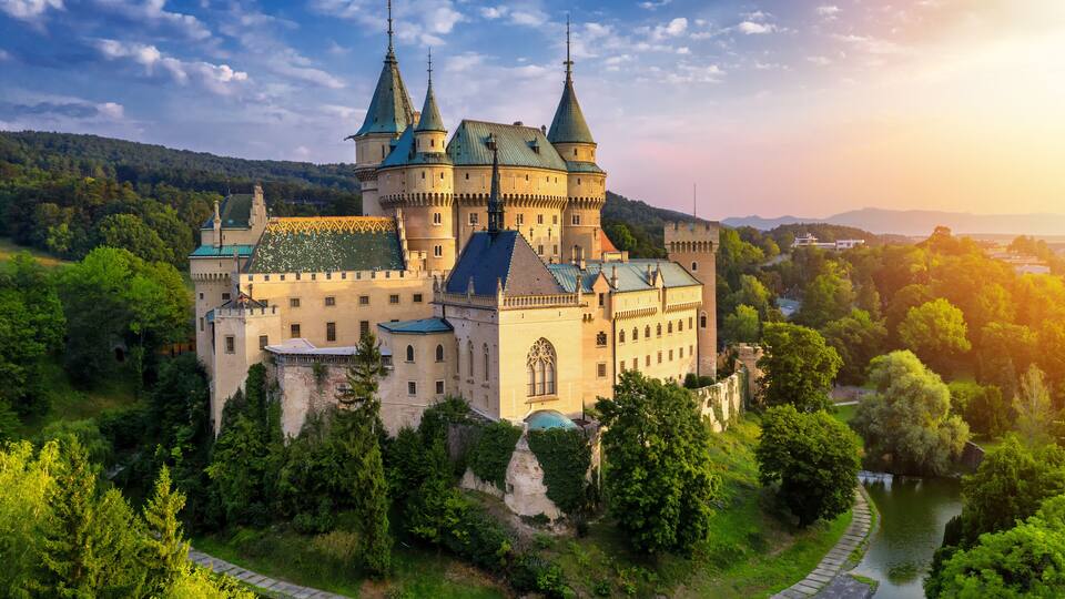 A castle with multiple towers, surrounded by lush greenery and a river.