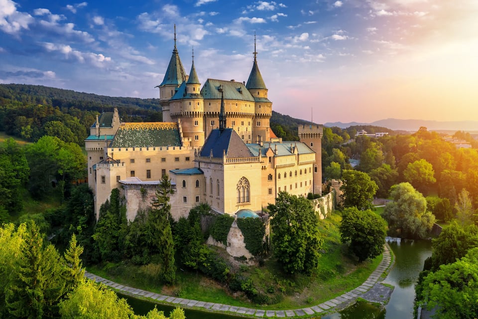 A castle with multiple towers, surrounded by lush greenery and a river.