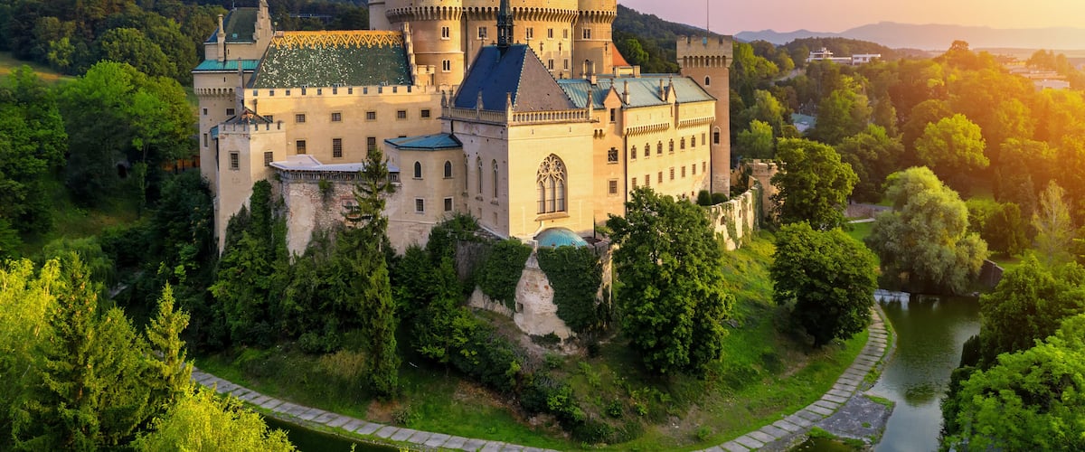 A castle with multiple towers, surrounded by lush greenery and a river.