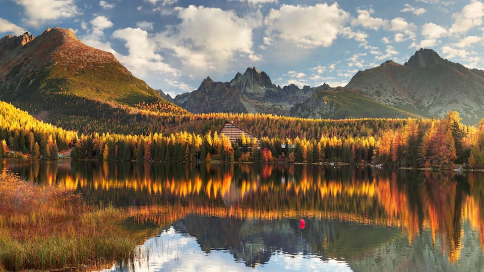 A mountain range with autumn foliage, a lake reflecting the scenery, and a bridge in the distance.