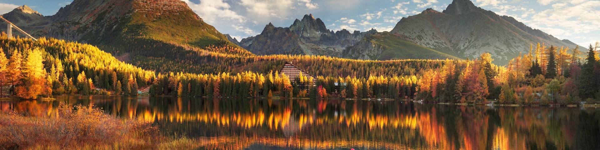 A mountain range with autumn foliage, a lake reflecting the scenery, and a bridge in the distance.