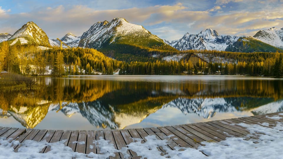 A serene lake with a wooden dock, surrounded by snow-capped mountains and dense forest.