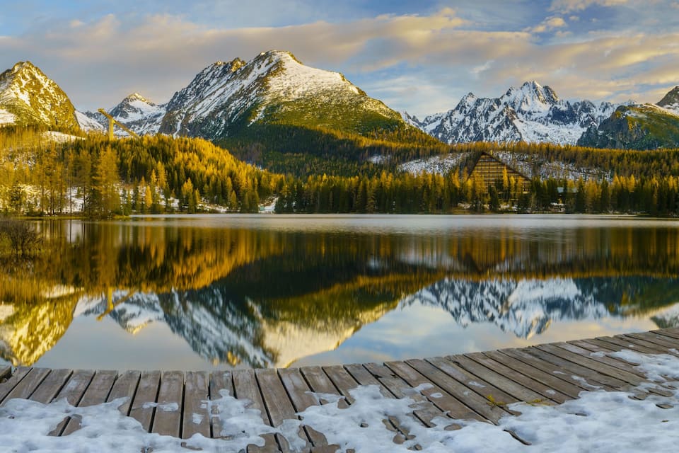 Un lago sereno con un muelle de madera, rodeado de montañas nevadas y un bosque denso.
