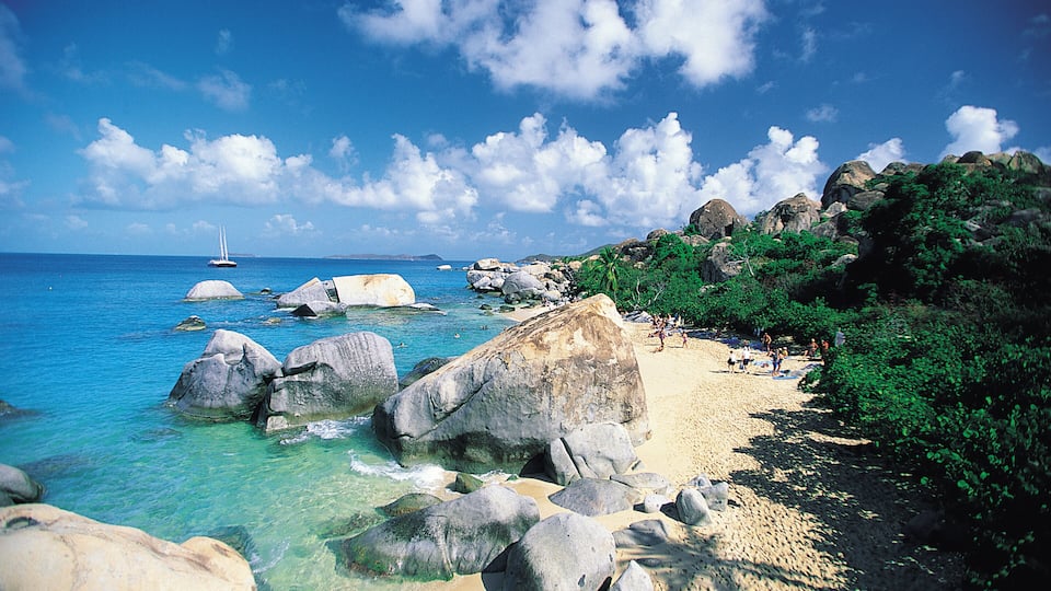 A rocky beach with clear turquoise water, large boulders, and a sailboat in the distance.