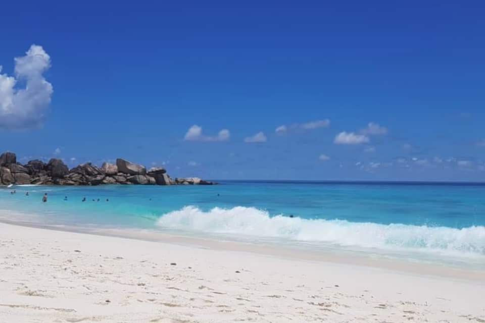 Una playa con agua turquesa cristalina, arena blanca y rocas grandes en la distancia.