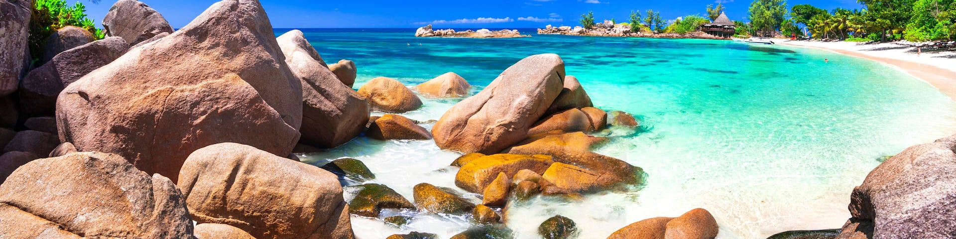Una playa tropical con rocas grandes, agua turquesa cristalina, una palmera y una cabaña con techo de paja.