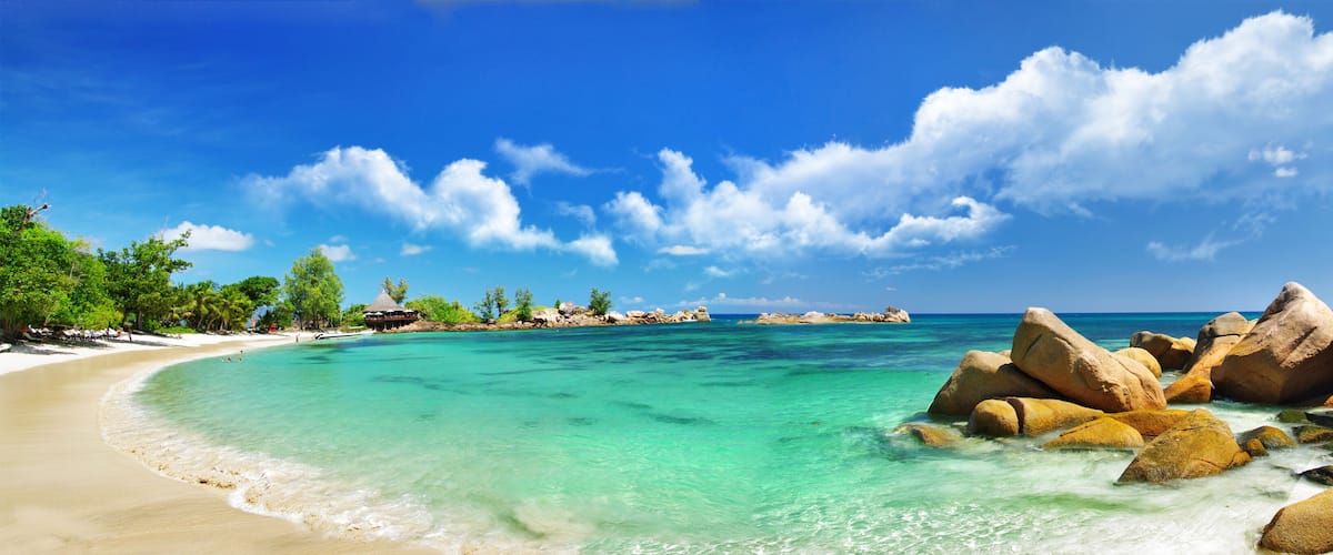 A tropical beach with clear turquoise waters, large rocks, and palm trees under a bright blue sky.