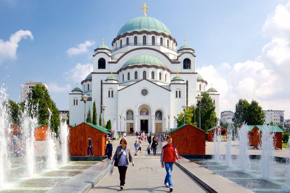 A large white church with green domes and a golden cross, surrounded by fountains and people walking.