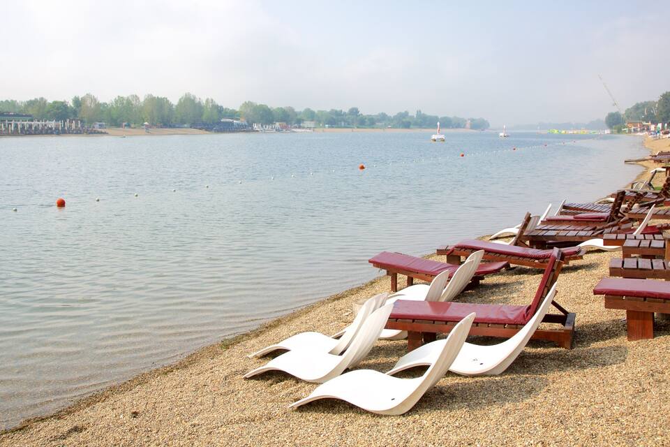 A lakeside beach with wooden lounge chairs and a row of boats on the water.