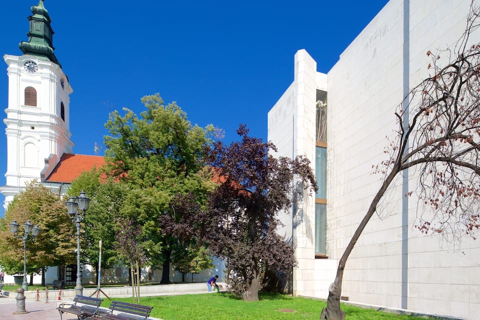Une église dotée d’un clocher, un bâtiment moderne et un parc avec des bancs et des arbres.