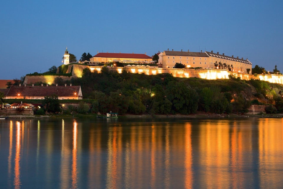 Un château historique perché sur une colline, surplombant une rivière paisible, avec des bâtiments illuminés et le clocher d’une église en arrière-plan.
