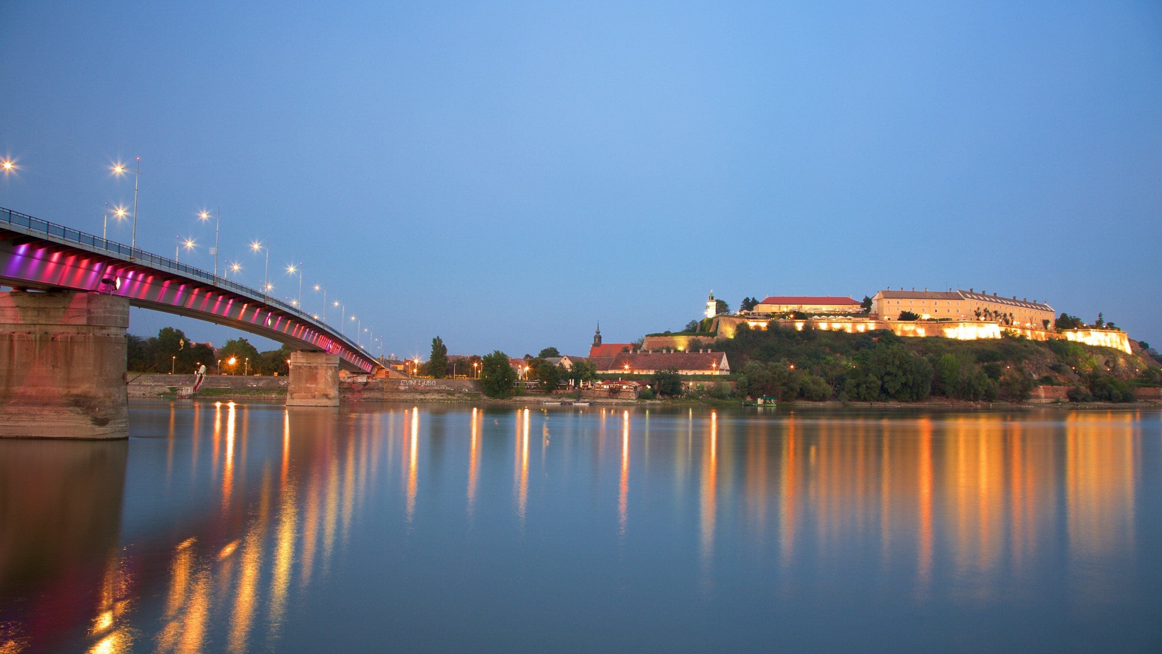 A bridge over a calm river with a castle on a hill in the background.