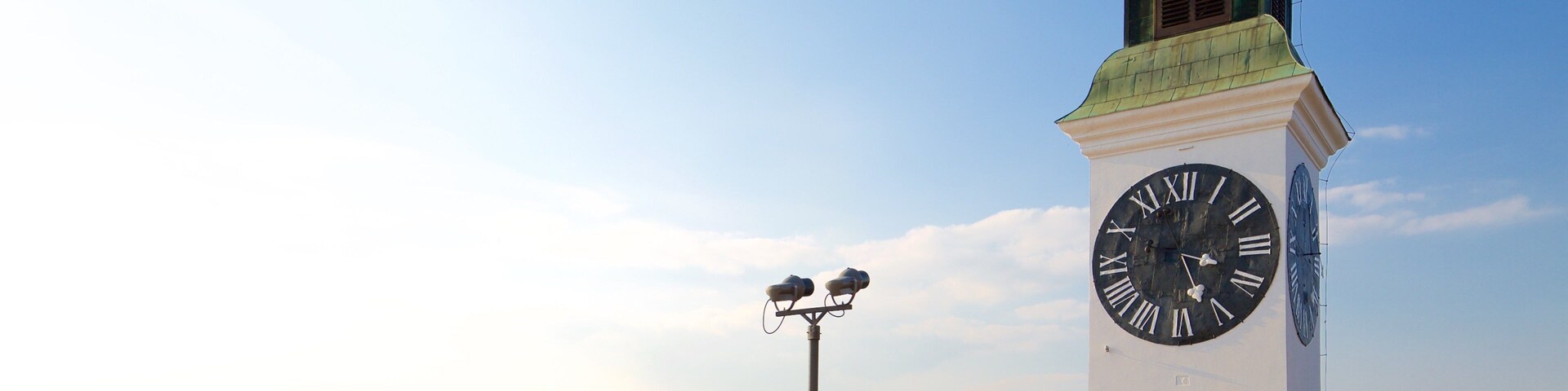 Um relógio de torre com telhado verde e um mostrador grande, cercado por um parque com pessoas e um rio ao fundo.