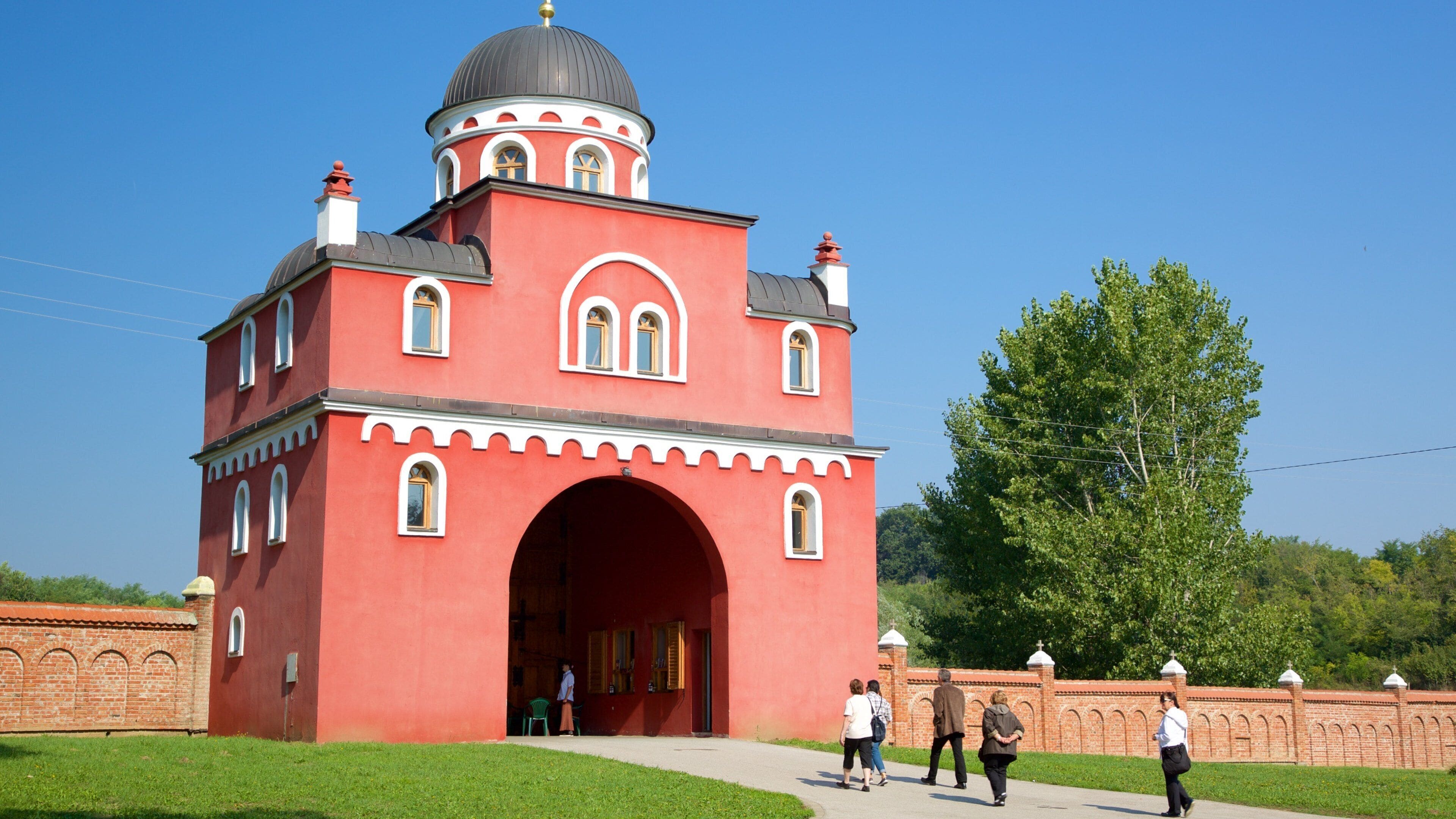 A red building with a dome and arched entrance, surrounded by a brick wall and greenery.