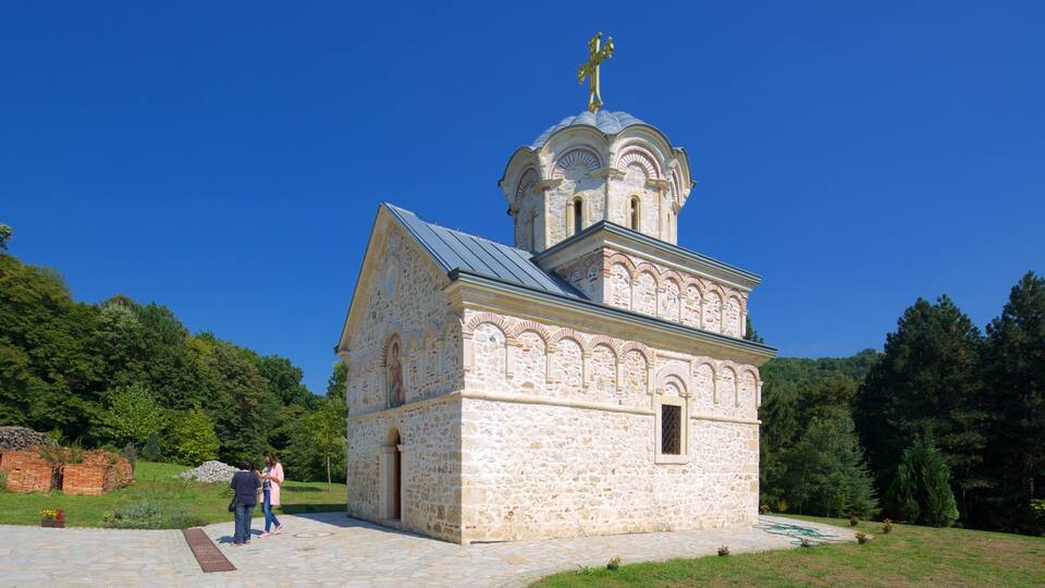 A small stone church with a golden cross on top, surrounded by greenery.