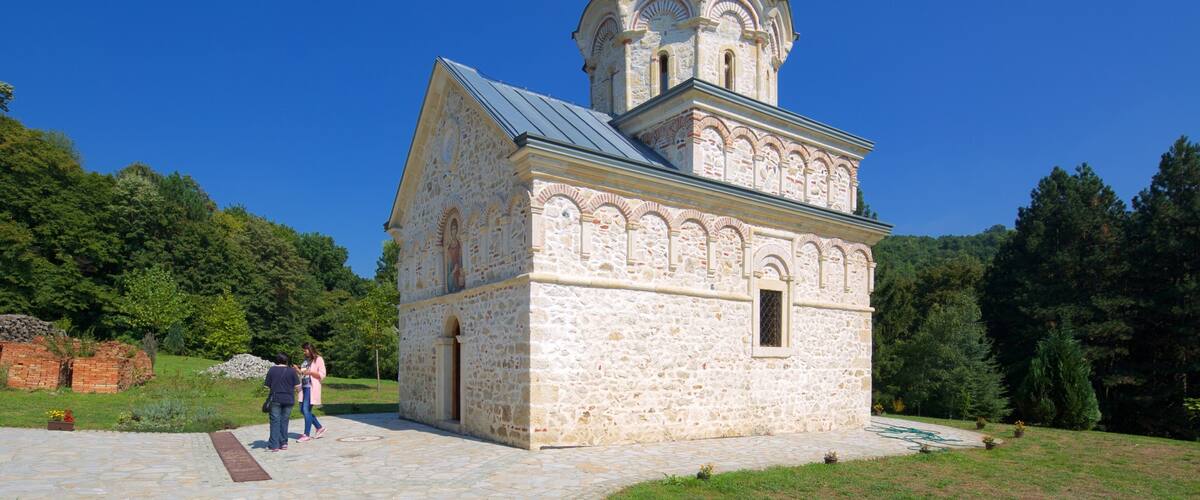 A small stone church with a golden cross on top, surrounded by greenery.