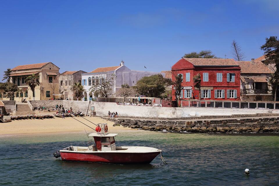 A coastal town with a red boat docked near the shore, buildings with red and blue facades, and a sandy beach with people in the background.