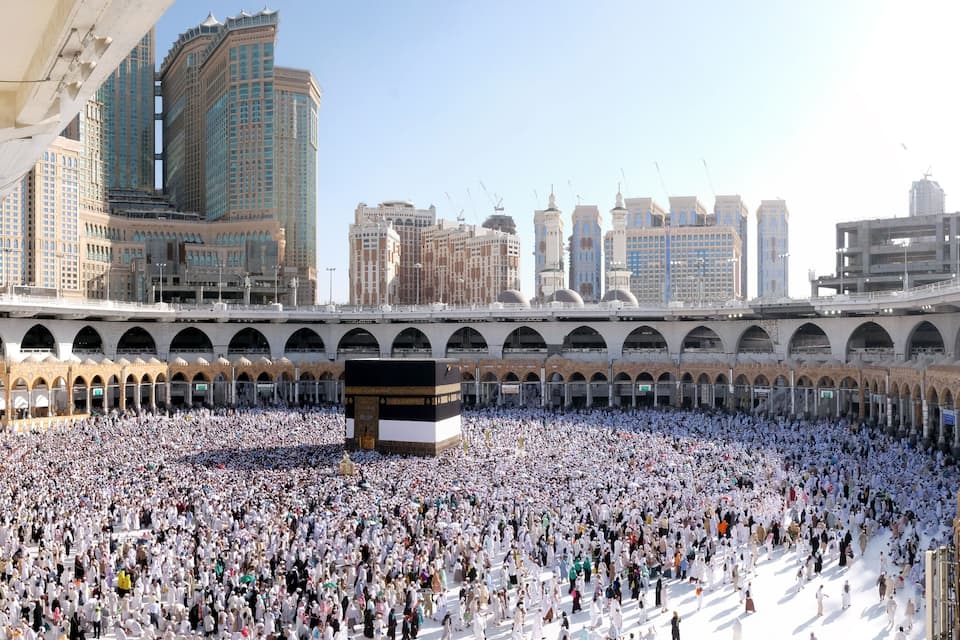 A large crowd surrounding the Kaaba in a historic courtyard with modern buildings in the background.