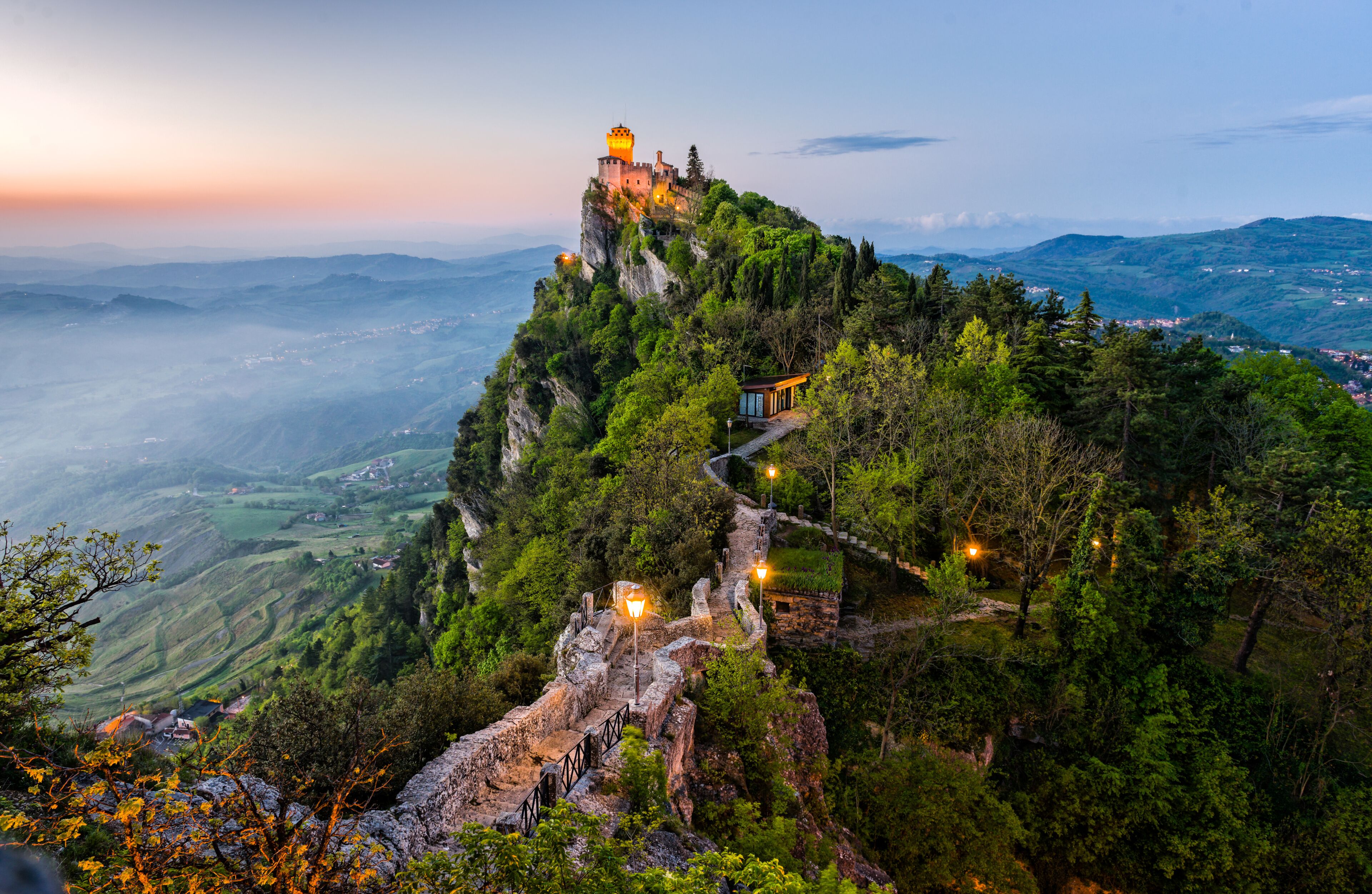 A castle on a mountain peak with a pathway leading up to it, surrounded by lush greenery and a valley below.