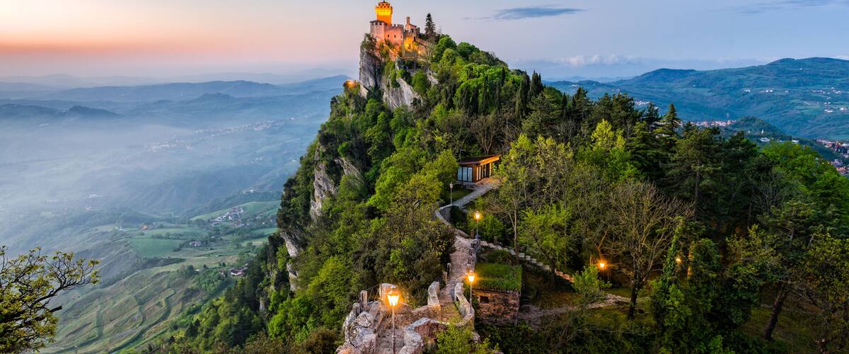 A castle on a mountain peak with a pathway leading up to it, surrounded by lush greenery and a valley below.