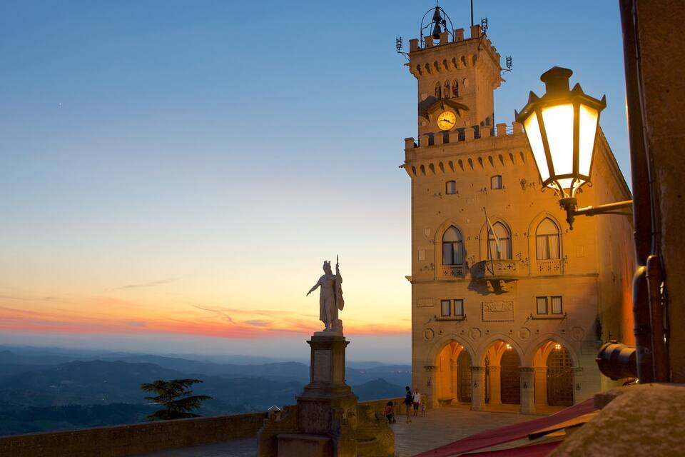 Ein Turm mit einer Statue davor, ein Sonnenuntergang und eine Bergkette im Hintergrund.