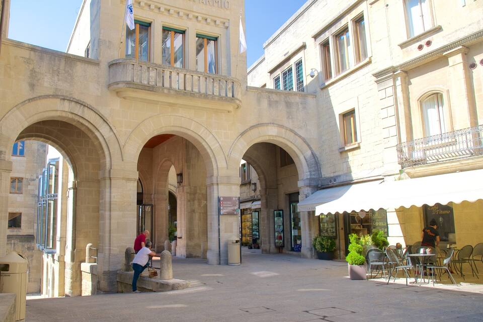 Ein historischer Platz mit steinernen Arkaden, einem Café und einer Person, die auf einer Bank sitzt.