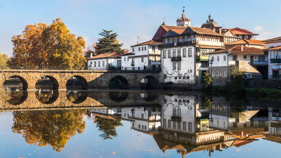 A stone bridge with arches over a calm river, reflecting buildings and trees.