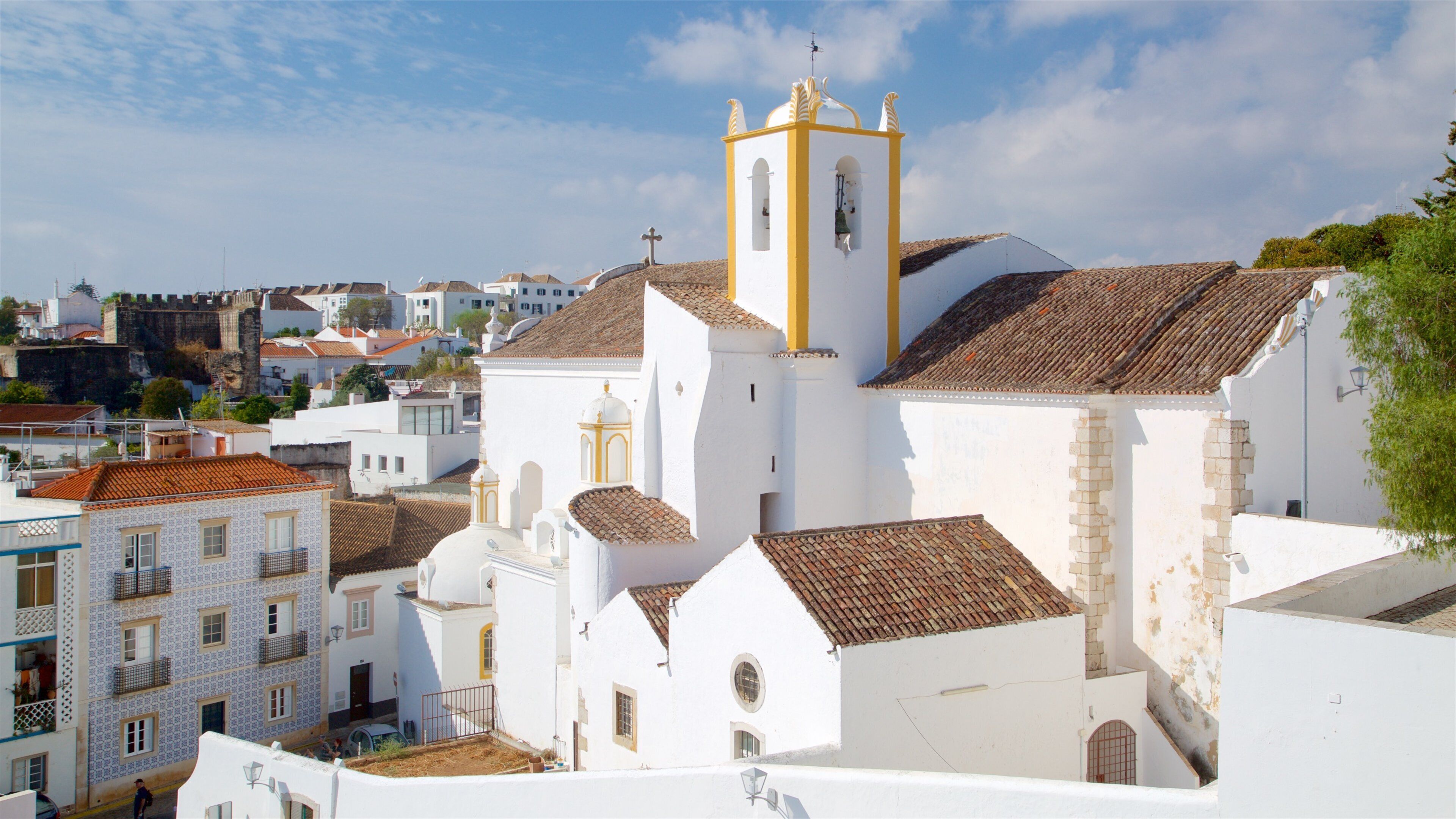 A white church with a yellow tower and a cross on top, surrounded by buildings with tiled roofs.