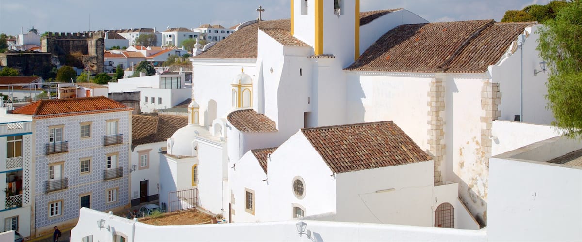A white church with a yellow tower and a cross on top, surrounded by buildings with tiled roofs.