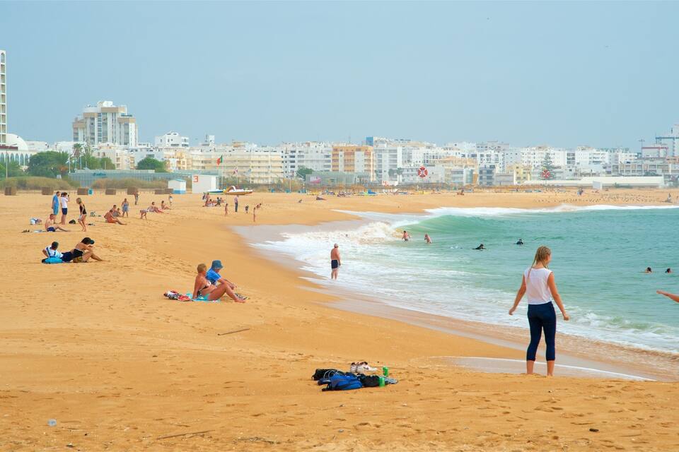 A sandy beach with people sunbathing and swimming. In the background, there are buildings and a clear sky.
