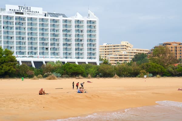 Une plage où l’on peut voir des gens se prélasser au soleil et jouer, un grand hôtel appelé TIVOLI MARINA VILLAMOURA, et d’autres bâtiments en arrière-plan.