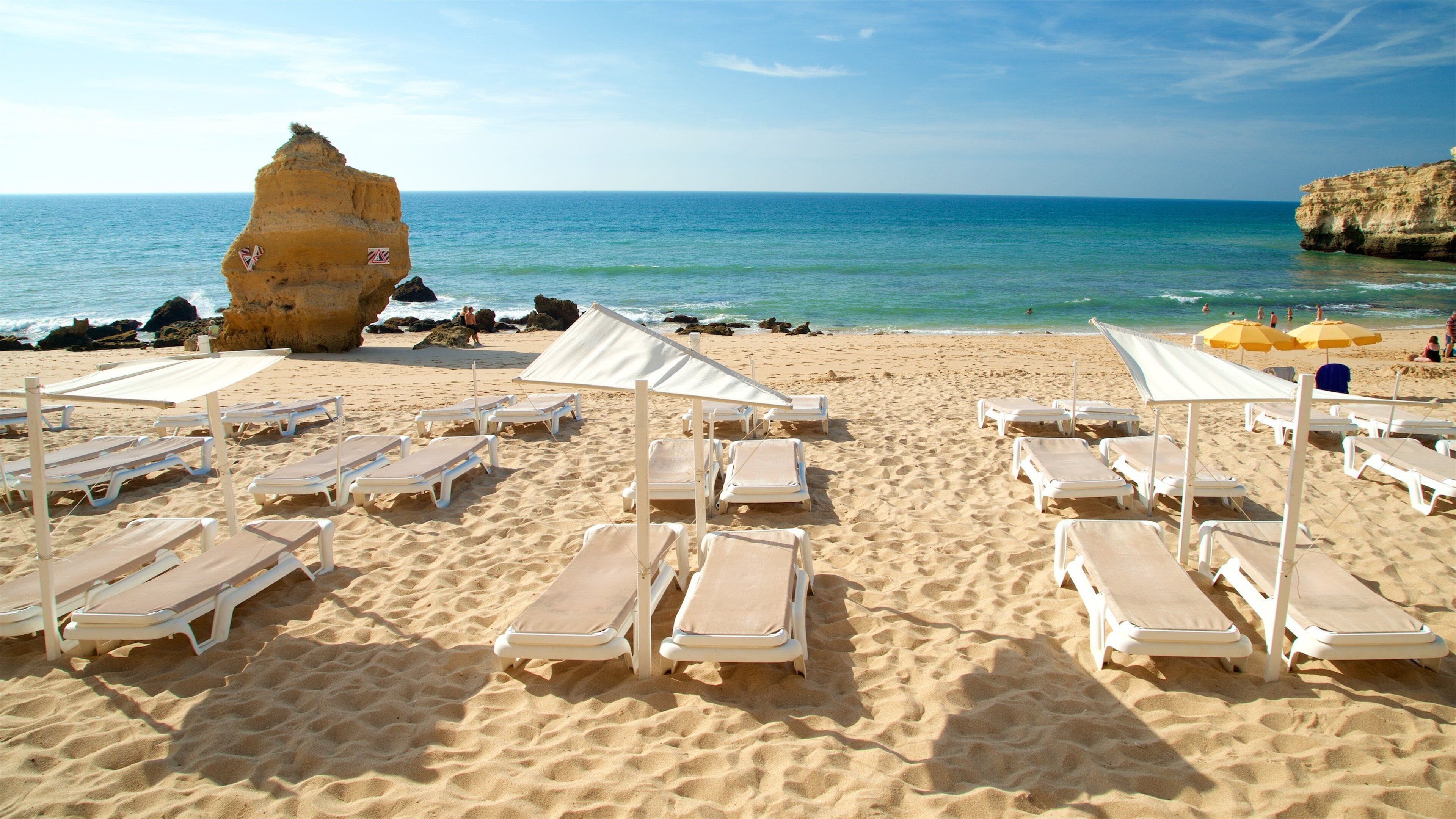 A beach with white sun loungers and umbrellas, a large rock formation, and the ocean.