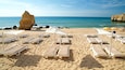 A beach with white sun loungers and umbrellas, a large rock formation, and the ocean.