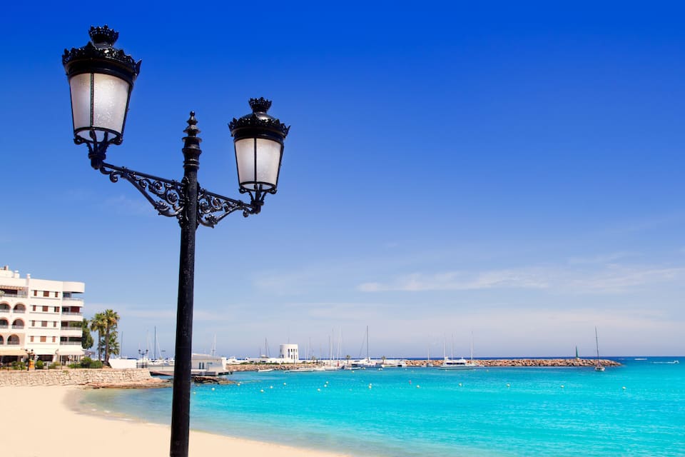 A beach with clear turquoise water, a white building, and a black street lamp.