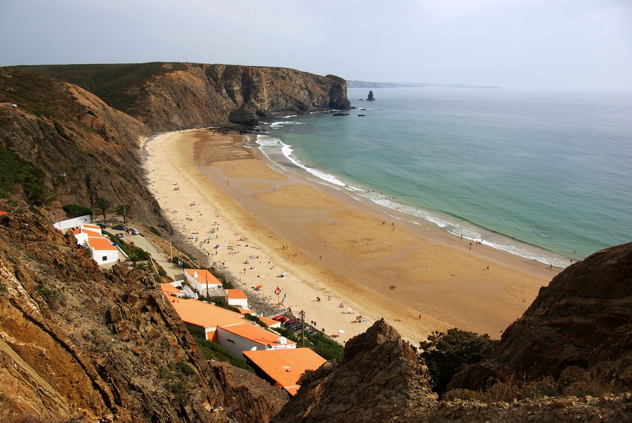 A coastal cliffside with a sandy beach, rocky terrain, and a lighthouse in the distance.