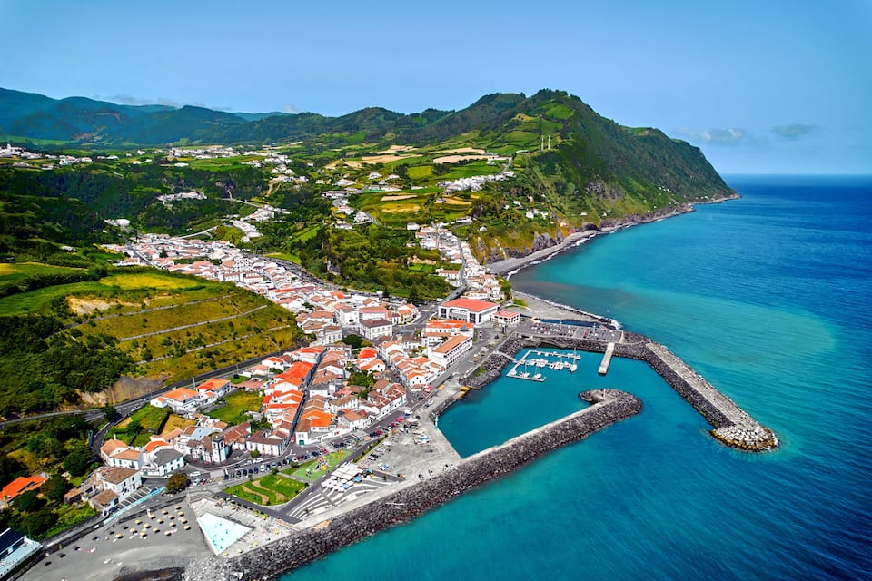 Un pueblo costero con un puerto, colinas verdes y un mar azul cristalino.