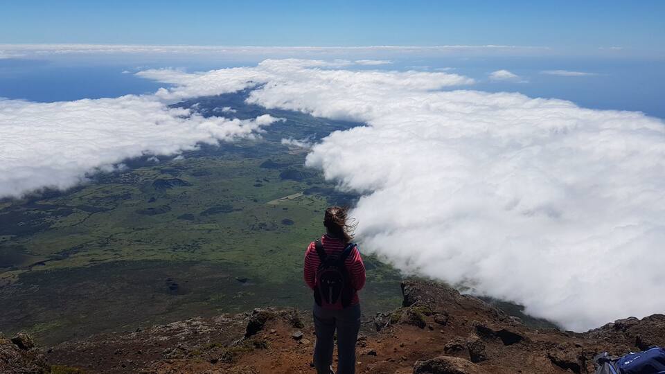 A person standing on a rocky mountain peak overlooking a valley of green fields and a sea of clouds.