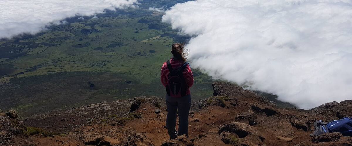 A person standing on a rocky mountain peak overlooking a valley of green fields and a sea of clouds.