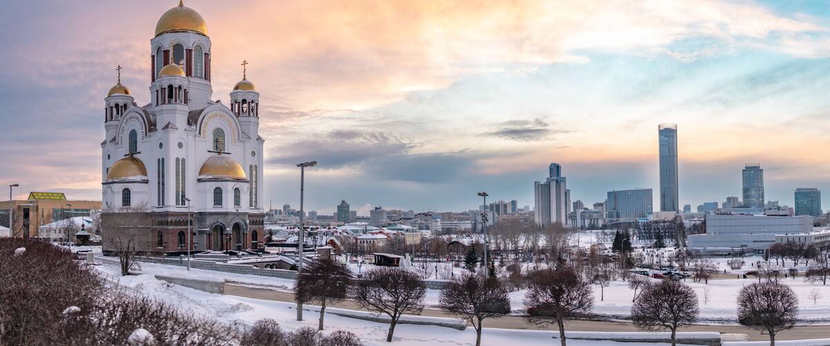 A cathedral with golden domes and a cross, surrounded by a snowy park and a city skyline with modern buildings.