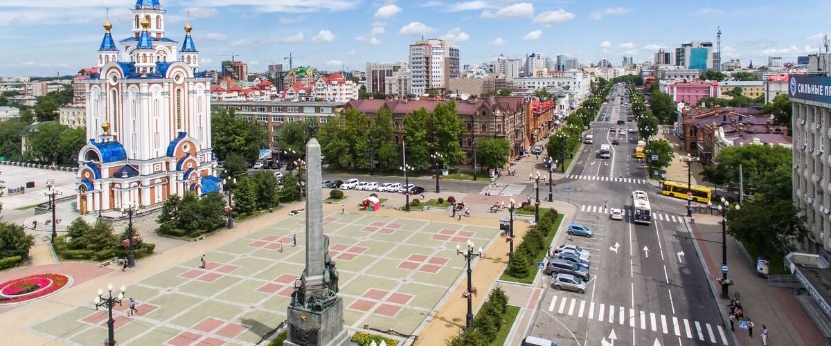 A cityscape with a prominent church with blue domes, a central monument, and a wide street lined with cars and pedestrians.