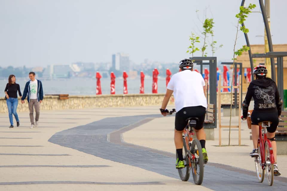 A waterfront promenade with cyclists, a couple walking, and a cityscape in the background.