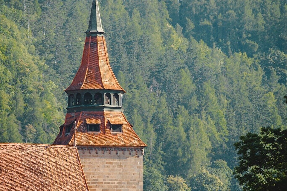 A church with a red-tiled roof and a tall spire, surrounded by a forested hillside.