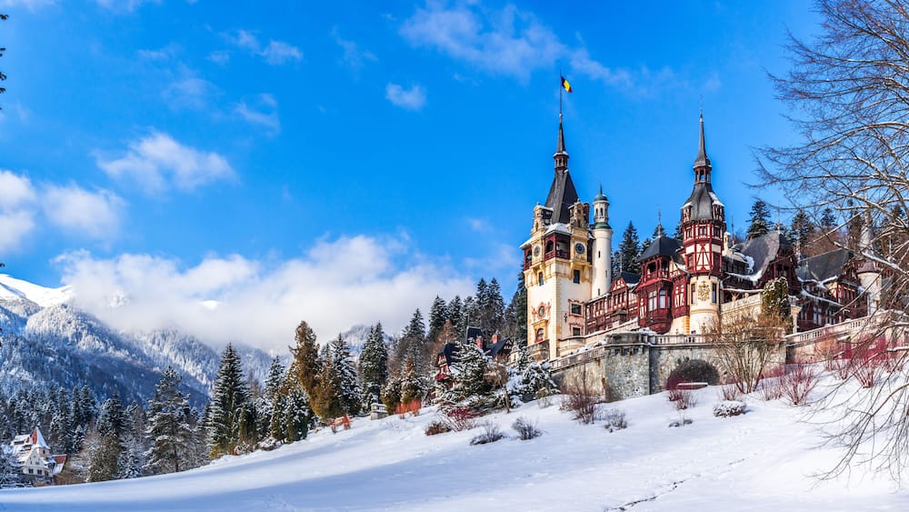 A snow-covered castle with two towers, surrounded by snow-laden trees and mountains.