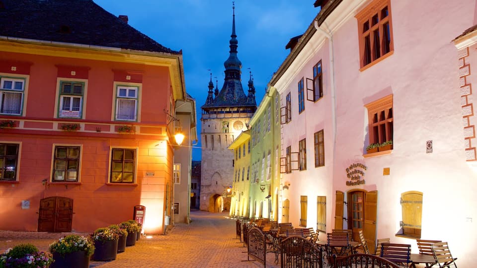 A cobblestone street lined with colorful buildings, a clock tower, and outdoor seating in the foreground.
