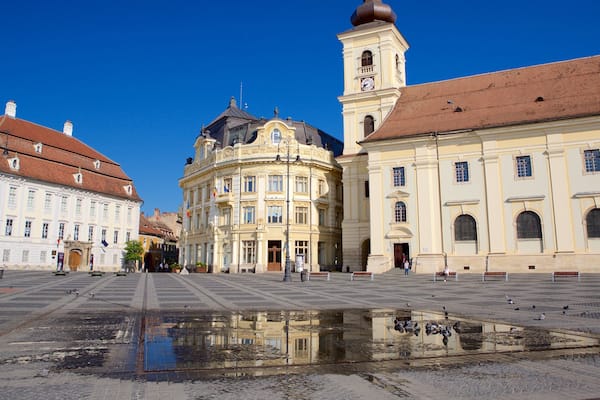 Ein historischer Platz mit einem zentralen Brunnen, umgeben von klassischer Architektur, darunter ein markanter Uhrturm.