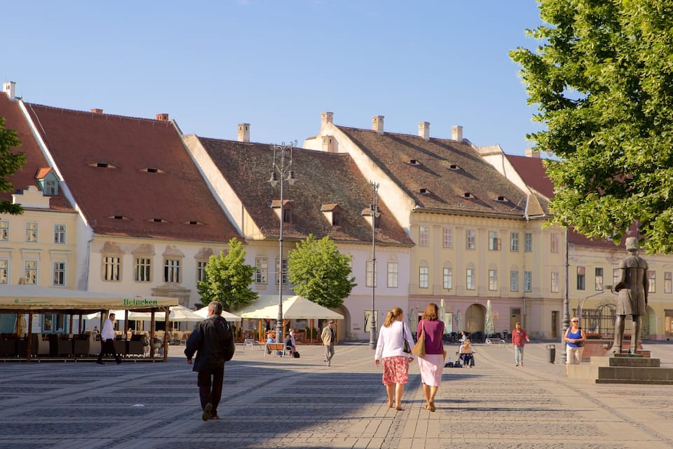 Uma praça histórica com edifícios tradicionais, uma estátua e pessoas caminhando.