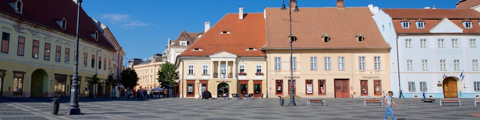 A square with cobblestone pavement, historic buildings with red and white facades, and a clear blue sky.