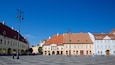 Una plaza con pavimento de adoquines, edificios históricos con fachadas rojas y blancas, y un cielo azul despejado.
