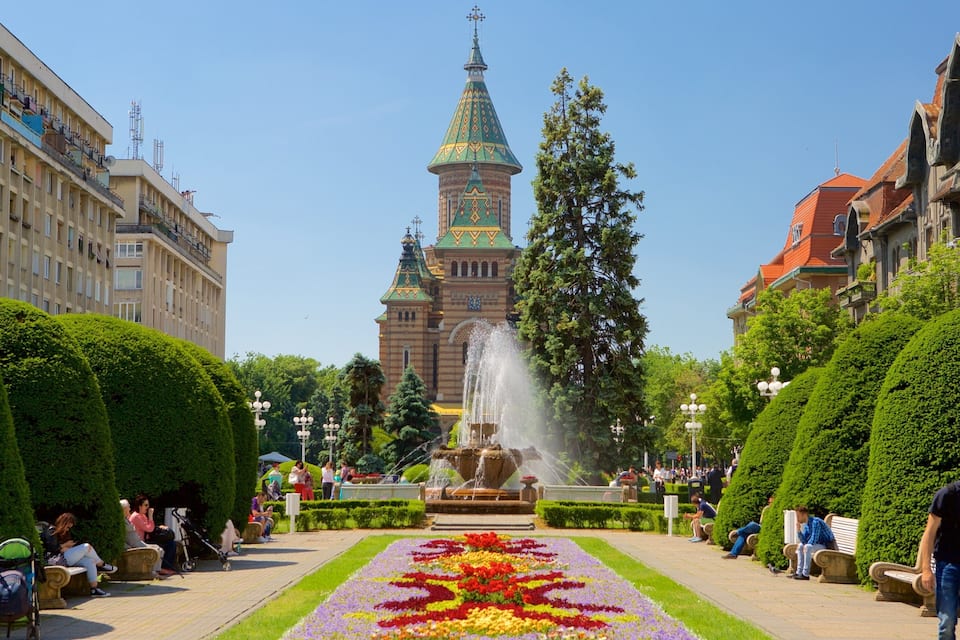 A fountain, flower beds, and a historic building with a green spire in the background.