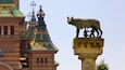 A statue of a bear on a pedestal with inscriptions, in front of a building with colorful domes and crosses.